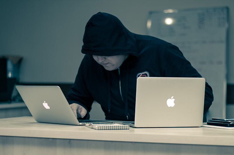Hacker working on two laptops wwaring a black hood